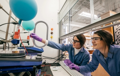 Two students wearing lab coats and protective goggles conduct an experiment with balloons and wiring.
